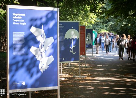 fotografia. wystawa Wyszehrad to ludzie na krakowskich plantach. ludzie przechadzają się pomiędzy plakatami ustawionymi w rzędzie wzdłuż alejki.